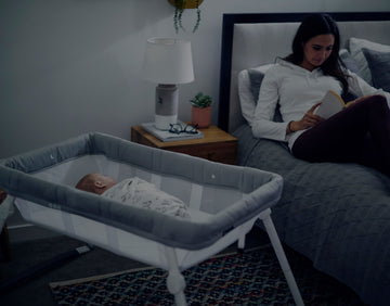 Swaddled baby in a Guava bassinet beside a woman reading while sitting up in bed